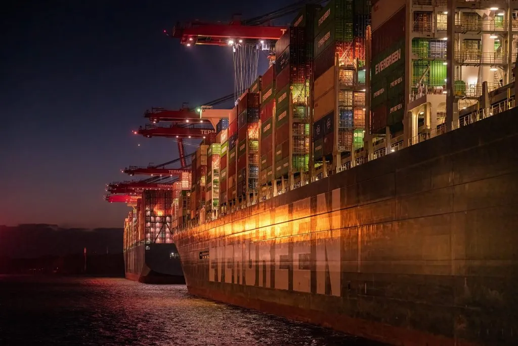 Large Evergreen cargo container ship docked at a port at night under bright industrial terminal lighting.