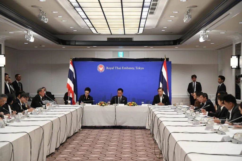 Prime Minister Anutin Charnvirakul chairs a meeting with Thai officials beneath a Royal Thai Embassy, Tokyo backdrop.