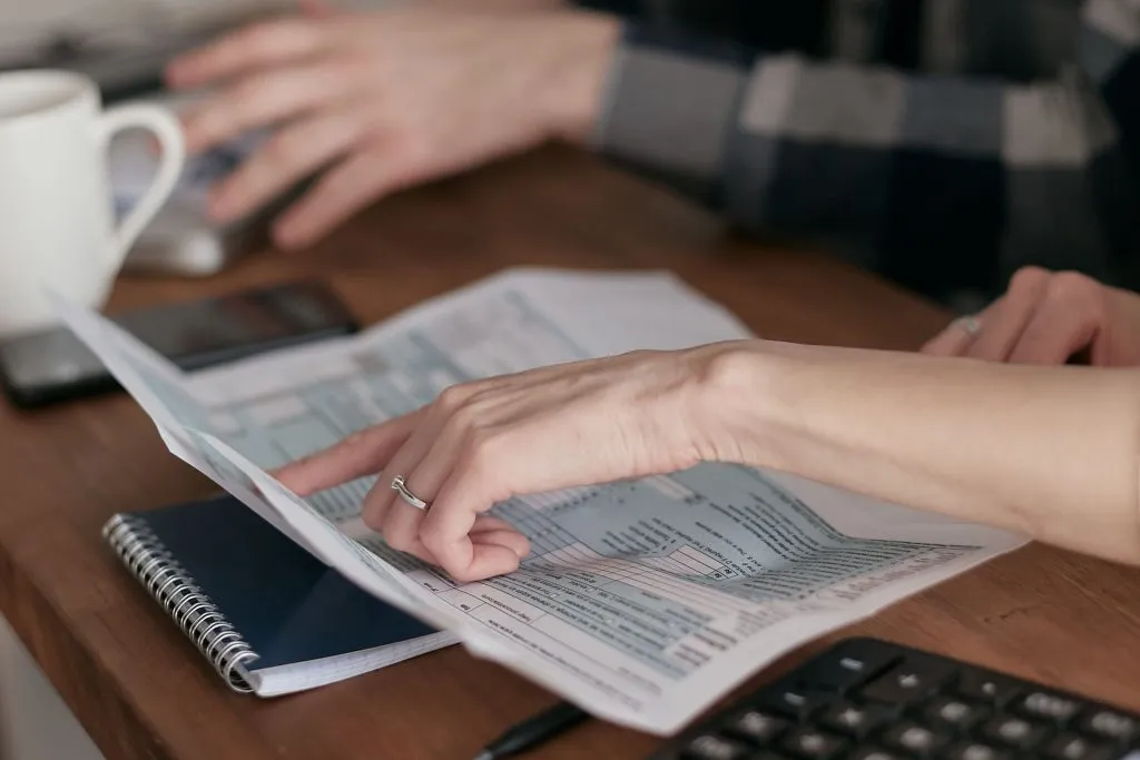 Hands examine documents beside a calculator on a wooden table.